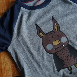Op de afbeelding: Een grijs en donkerblauw longsleeve shirt met een cartoon vleermuis afbeelding. De vleermuis is bruin met gele accenten en heeft een tandenrij.