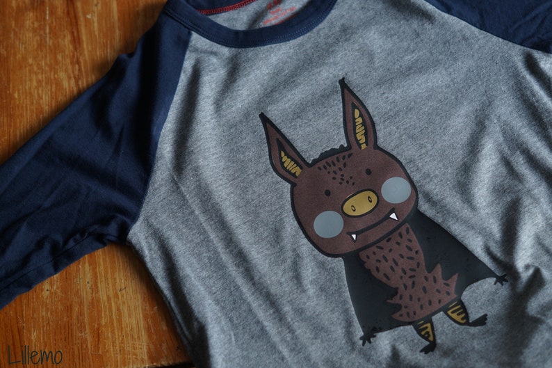 Op de afbeelding: Een grijs en donkerblauw longsleeve shirt met een cartoon vleermuis afbeelding. De vleermuis is bruin met gele accenten en heeft een tandenrij.