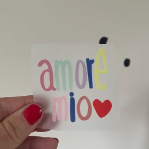 Könnte beinhalten: Ein farbenfroher Aufkleber mit den Worten "amore mio" in verschiedenen Pastellfarben, mit einem roten Herz, das das Wort "Liebe" ersetzt. Der Aufkleber wird von einer Hand mit rotem Nagellack gehalten.