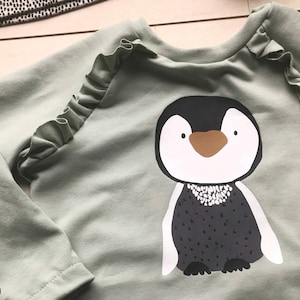 May include: A sage green long-sleeved shirt with a ruffle detail on the shoulders. The shirt features a graphic print of a penguin with a black and white body and a brown beak.