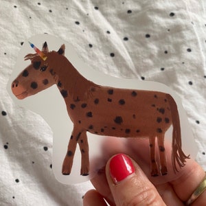 Könnte beinhalten: Ein brauner und schwarz gefleckter Einhorn-Aufkleber mit einem Regenbogenhorn. Der Aufkleber befindet sich auf einem weißen Hintergrund mit schwarzen Punkten.