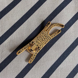 Pode incluir: Um patch bordado dourado em forma de leopardo estilizado, deitado com as patas estendidas. O patch tem uma aparência texturizada e está sobre um fundo listrado.