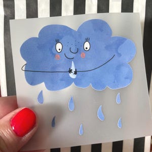 Könnte beinhalten: Ein blauer Wolkenaufkleber mit einem lächelnden Gesicht, rosigen Wangen und Regentropfen. Die Wolke hat ein niedliches Design mit einem fröhlichen Ausdruck. Der Aufkleber befindet sich auf einem durchscheinenden Hintergrund.