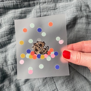 Może przedstawiać: Ilustracja brązowego i białego niedźwiedzia otoczonego kolorowymi kropkami konfetti na przezroczystym tle.