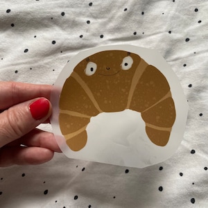 Könnte beinhalten: Ein braunes Cartoon-Croissant mit einem lächelnden Gesicht und weißen Augen. Das Croissant befindet sich auf einem weißen Hintergrund mit schwarzen Punkten.