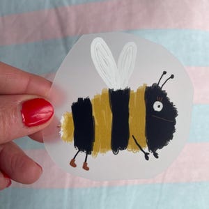 Peut inclure: Autocollant fantaisiste représentant une abeille de dessin animé avec un corps rayé noir et jaune, des ailes blanches et de petites bottes marron. L'abeille a une expression amicale avec un grand œil et des antennes. L'autocollant est sur fond transparent.