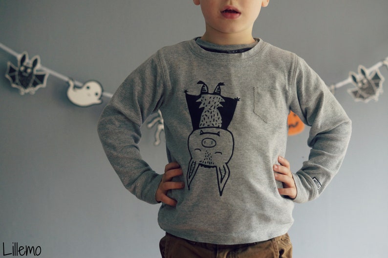 Op de afbeelding: Een grijs sweatshirt met een zwart-witte illustratie van een vleermuis die ondersteboven hangt. De vleermuis heeft een glimlachend gezicht en een kleine neus.