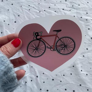 Può includere: Un adesivo a forma di cuore rosa con un'illustrazione di bicicletta rossa.