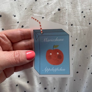 Könnte beinhalten: Ein blauer und weißer Saftkarton mit einem roten und weißen Strohhalm. Der Saftkarton hat einen lächelnden roten Apfel mit einem grünen Blatt auf der Vorderseite und den Text "Mariecchens Äppelsäftchen".