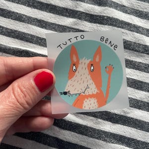 Könnte beinhalten: Ein runder Aufkleber mit einem Cartoon-Fuchs in Orange und Weiß auf türkisblauem Hintergrund. Der italienische Ausdruck "TUTTO BENE" ist in Schwarz geschrieben. Der Fuchs macht eine "Okay"-Handgeste.