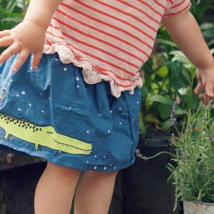 May include: A young child wearing a blue skirt with a green alligator print and white stars. The skirt has a red and white striped shirt underneath. The child is reaching out to touch a lavender plant.