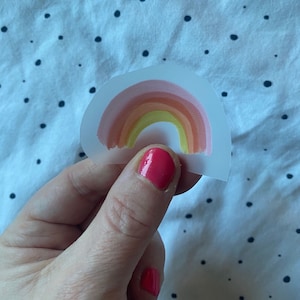 Può includere: Un adesivo trasparente con un design arcobaleno. L'arcobaleno ha sfumature di rosa, arancio, giallo e verde.