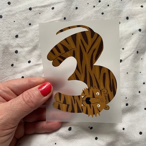 Könnte beinhalten: Ein brauner und orangefarbener Tiger in Form der Zahl drei. Der Tiger hat schwarze Streifen und ein weißes Gesicht mit schwarzen Augen und einer schwarzen Nase.