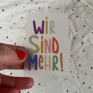 Könnte beinhalten: Ein bunter Sticker mit dem Text "Wir sind mehr!" Die Buchstaben sind in verschiedenen Farben, darunter Lila, Grün, Orange, Rosa und Blau.