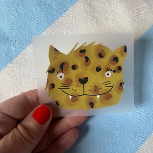 Könnte beinhalten: Ein gelber und brauner Leoparden-Print-Aufbügel-Patch mit einem lächelnden Gesicht und schwarzen Wimpern.