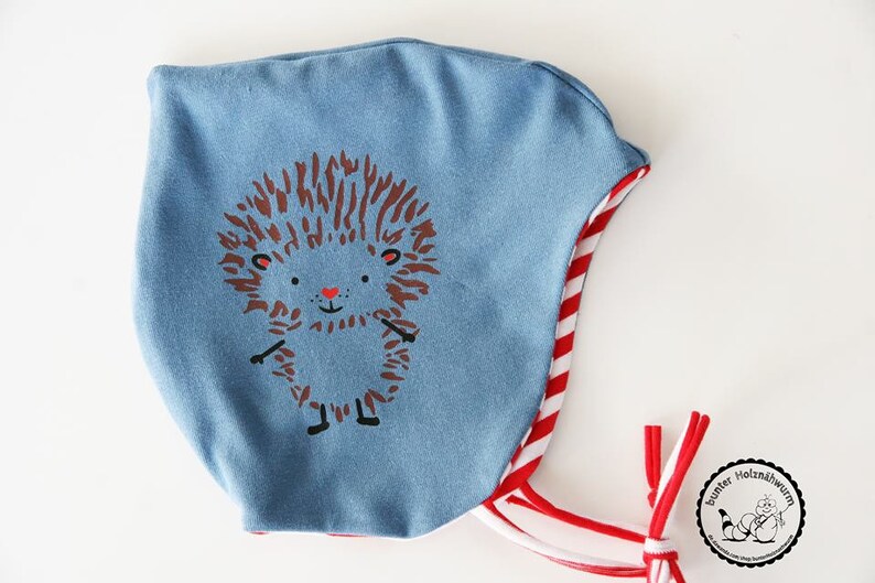 May include: A light blue baby bonnet featuring a cartoon hedgehog design. The bonnet has a red and white striped trim and ties. The hedgehog has brown quills, black feet, and a red heart-shaped nose.