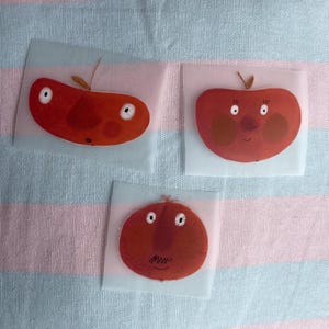Op de afbeelding: Drie rode appelvormige illustraties met verschillende gezichtsuitdrukkingen. De appels bevinden zich op een witte en roze gestreepte achtergrond.