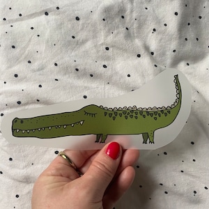 May include: A green alligator sticker with closed eyes and a toothy grin. The sticker is on a white background with black polka dots.