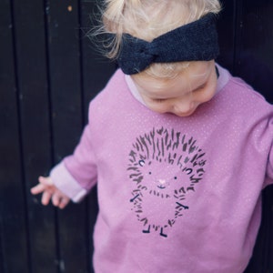 May include: A child's outfit featuring a light purple sweatshirt with a hedgehog graphic and a dark grey headband. The sweatshirt has a round neck and long sleeves. The image is set against a dark wooden background.