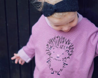 Könnte beinhalten: Ein Kinder-Outfit mit einem hellvioletten Sweatshirt mit einem Igel-Motiv und einem dunkelgrauen Stirnband. Das Sweatshirt hat einen Rundhalsausschnitt und lange Ärmel. Das Bild ist vor einem dunklen Holzhintergrund aufgenommen.