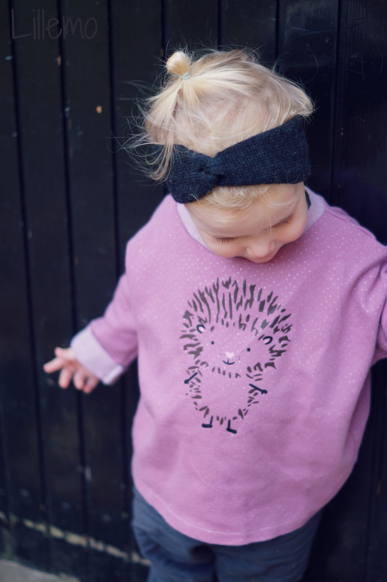 May include: A child's outfit featuring a light purple sweatshirt with a hedgehog graphic and a dark grey headband. The sweatshirt has a round neck and long sleeves. The image is set against a dark wooden background.