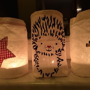 May include: A collection of illuminated fabric lanterns. One has a red and white gingham star, another a hedgehog design, and a third shows a child in a red and white gingham coat. The lanterns are displayed on a white surface.