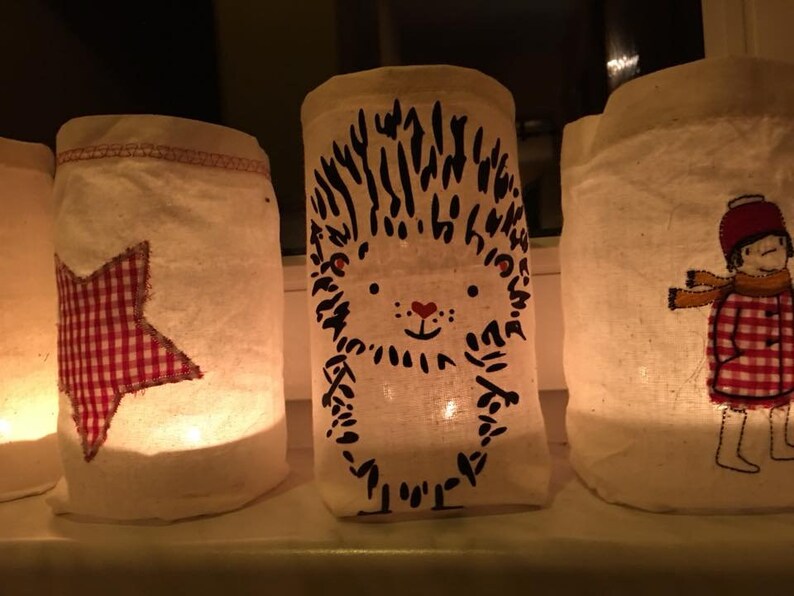 May include: A collection of illuminated fabric lanterns. One has a red and white gingham star, another a hedgehog design, and a third shows a child in a red and white gingham coat. The lanterns are displayed on a white surface.