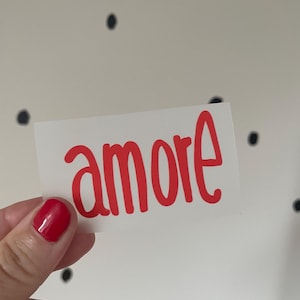 Könnte beinhalten: Ein weißer rechteckiger Aufkleber mit dem Wort "amore" in roter, kursiver Schrift. Der Aufkleber wird von einer Hand mit rotem Nagellack gehalten. Der Hintergrund ist eine weiße Wand mit schwarzen Punkten.