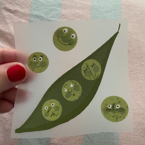 Puede incluir: Una ilustración caprichosa de una vaina de guisantes verdes con cinco guisantes, cada uno con una expresión facial única. Los guisantes muestran una gama de emociones, desde sonrientes hasta somnolientos, todo en tonos verdes. La obra de arte está sobre papel translúcido.