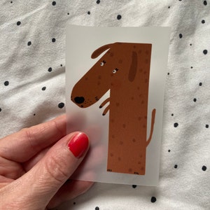 Könnte beinhalten: Ein brauner, hundeförmiger Aufkleber mit schwarzen Augen und einer schwarzen Nase. Der Aufkleber befindet sich auf einem transparenten Hintergrund.