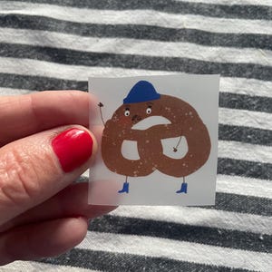 Könnte beinhalten: Ein skurriler Aufkleber mit einer Cartoon-Brezel-Figur. Die Brezel ist braun, trägt einen blauen Hut und Stiefel und hat einen überraschten Gesichtsausdruck. Der Aufkleber hat einen transparenten Hintergrund.