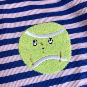 Könnte beinhalten: Ein gerippter Stoff mit horizontalen Streifen in Dunkelblau und Hellrosa. In der Mitte befindet sich ein flauschiger, kreisförmiger, lindgrüner Tennisball-Aufnäher mit einem gestickten Gesicht, bestehend aus zwei Augen, einer kleinen Nase und einem Mund.
