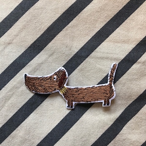 Könnte beinhalten: Brauner gestickter Dackel-Aufnäher mit gelbem Halsband. Der Aufnäher befindet sich auf einem gestreiften Stoffhintergrund.