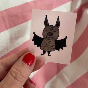 Könnte beinhalten: Eine braune und schwarze Cartoon-Fledermaus mit geschlossenen Augen und einem zahnigen Grinsen. Die Fledermaus befindet sich auf einem rosa Hintergrund.