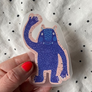 Könnte beinhalten: Ein blauer Monster-Aufkleber mit einem zahnigen Grinsen und einem erhobenen Arm, der Hallo winkt. Der Aufkleber befindet sich auf einem rosa und weißen Tupfenhintergrund.