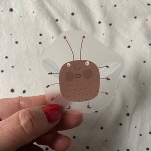 Op de afbeelding: Een bruine kever met een wit gezicht en vleugels, op een doorzichtige sticker.