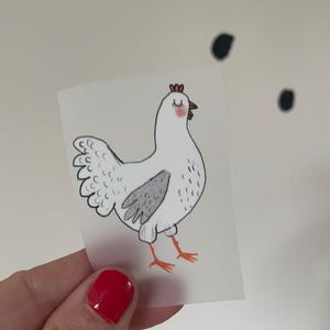 Könnte beinhalten: Illustration eines weißen Huhns mit einem grauen Flügel und roten Akzenten. Das Huhn hat geschlossene Augen und einen roten Kamm. Das Bild wird von einer Hand mit rotem Nagellack gehalten.
