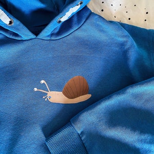 Puede incluir: Sudadera con capucha azul con un diseño de caracol de dibujos animados. El caracol tiene una concha marrón y es de color beige claro. La sudadera tiene una capucha con cordón y puños acanalados.
