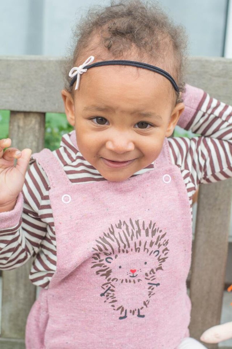 May include: A child wearing a pink dungaree dress featuring a hedgehog design, paired with a striped top. The child has a black headband with a white bow.