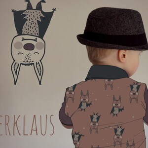 Op de afbeelding: Een jong kind draagt een bruine hoed met een zwarte band en een bruin shirt met een patroon van vleermuizen. Het shirt heeft de tekst "Flederklaus" en "Lillemo" erop. Het kind staat voor een muur met een tekening van een vleermuis.