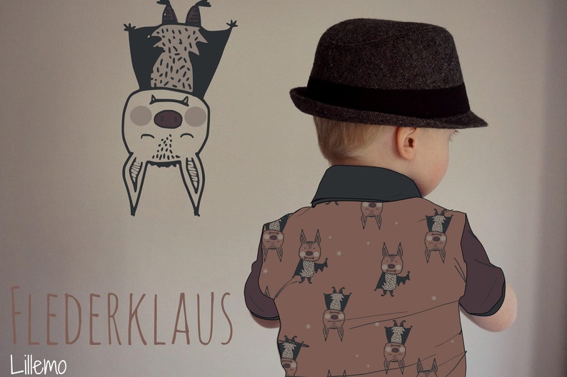 Op de afbeelding: Een jong kind draagt een bruine hoed met een zwarte band en een bruin shirt met een patroon van vleermuizen. Het shirt heeft de tekst "Flederklaus" en "Lillemo" erop. Het kind staat voor een muur met een tekening van een vleermuis.