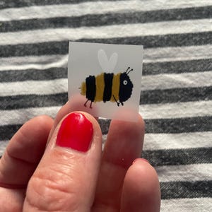 Könnte beinhalten: Ein kleiner, quadratischer Aufkleber mit einer Cartoon-Biene mit schwarzem Körper, gelben Streifen und weißen Flügeln. Die Biene hat ein skurriles Design mit einem Auge und kleinen Beinen. Der Aufkleber wird von einer Person gehalten.