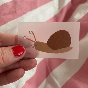 Könnte beinhalten: Eine Cartoon-Schnecke mit einer braunen Schale und einem hellbraunen Körper. Die Schnecke hat drei Fühler mit blauen Augen an den Enden.