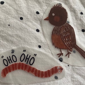 Könnte beinhalten: Zwei transparente Aufkleber aus Kunststoff mit Illustrationen. Ein Aufkleber zeigt einen braunen Vogel mit schwarzem Schnabel und schwarzen Augen. Der andere Aufkleber zeigt einen roten Wurm mit dem Text "ÖHO ÖHO" in Schwarz.