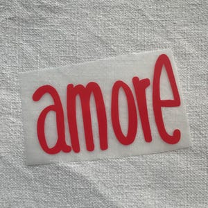 Peut inclure: Un autocollant en vinyle rouge avec le mot "amore" écrit en cursive. L'autocollant est sur un support blanc et est présenté sur un fond de tissu texturé blanc cassé.