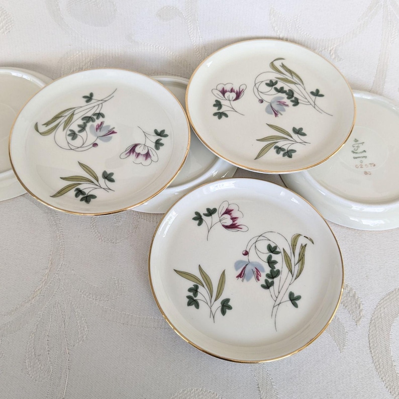 May include: Set of small, white ceramic plates with a gold rim. Each plate features a floral design with purple, blue, and green accents. The plates are arranged on a white surface, showcasing their delicate details and vintage aesthetic.