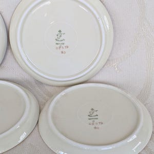 May include: A collection of off-white porcelain saucers. Each saucer has a slightly raised rim and a decorative emblem on the underside, with the text "Germany" and other markings. The saucers are arranged on a light-colored surface.