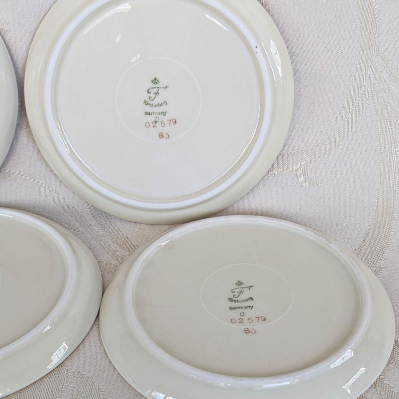 May include: A collection of off-white porcelain saucers. Each saucer has a slightly raised rim and a decorative emblem on the underside, with the text "Germany" and other markings. The saucers are arranged on a light-colored surface.