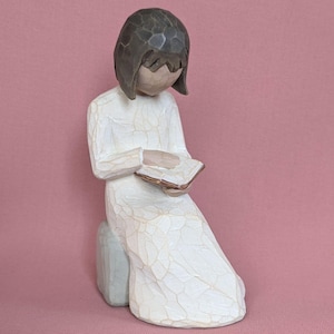 Könnte beinhalten: Eine geschnitzte Figur einer Person in einem langen weißen Kleid, die sitzt und ein Buch liest. Die Figur hat kurze braune Haare und kniet auf einem kleinen, hellfarbigen Block. Das Buch hat goldfarbene Kanten. Der Hintergrund ist einfarbig rosa.