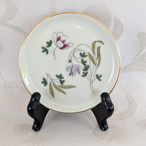 May include: A small, off-white ceramic plate with a gold rim, featuring a floral design in purple, blue, and green. The plate is displayed on a black stand, showcasing its decorative pattern. This vintage plate is perfect for serving small portions.